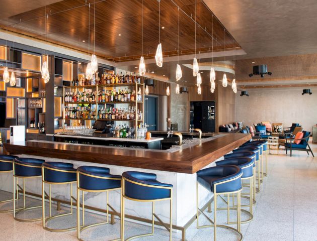 The image shows a modern bar with a wooden countertop, blue bar stools, hanging lights, and a well-stocked display of bottles.