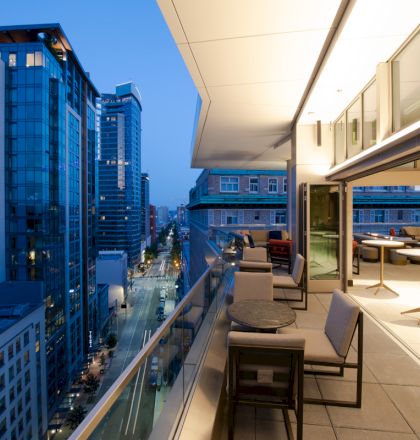 A modern cityscape at dusk, viewed from a balcony with comfortable seating and tables overlooking a lit urban street and tall buildings.