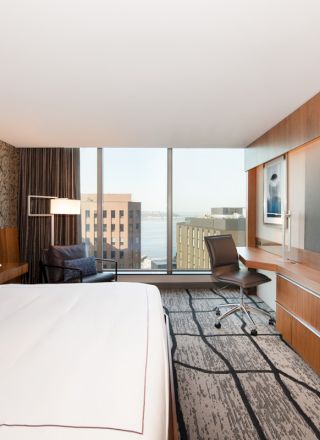 A modern hotel room with a large bed, a wall-mounted TV, a desk, a chair, and a window view of city buildings and water.