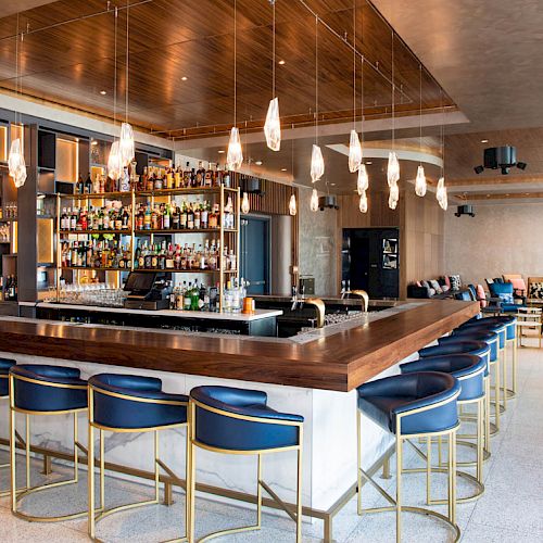 The image shows a modern bar setup with a central counter, blue bar stools, hanging lights, and a stocked bar in the background.