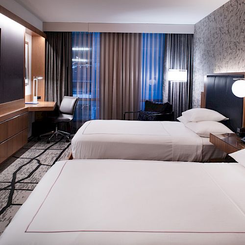 A hotel room with two beds, a flat-screen TV, a desk with a chair by the window, and modern decor, including patterned wallpaper and lamps.