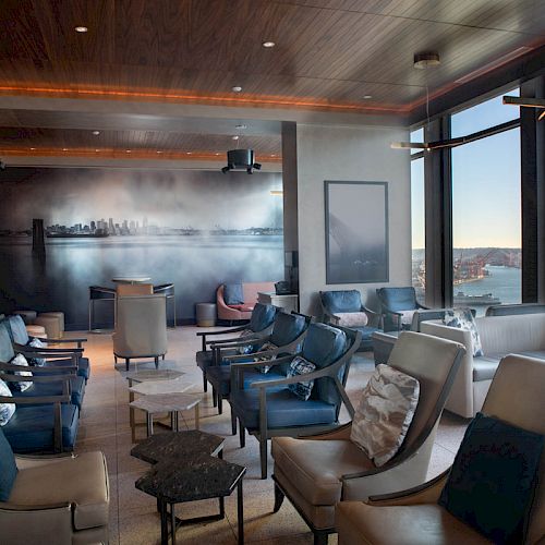 A modern lounge with various chairs and tables, large windows with an ocean view, a wall mural, and serene, ambient lighting.
