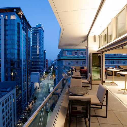 A modern balcony with tables and chairs, overlooking a city street lined with tall buildings at dusk.