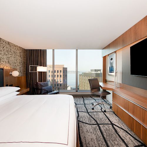 The image shows a modern hotel room with a large bed, a desk with a chair, a large TV, and a window offering a city view.