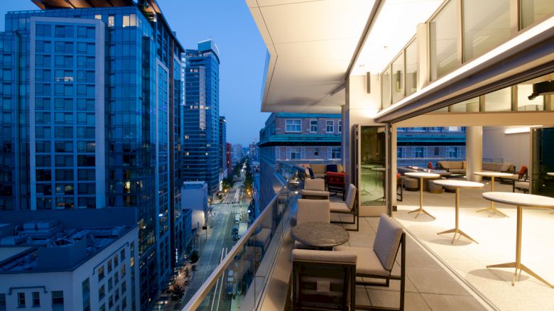 The image depicts a modern rooftop terrace overlooking a cityscape at dusk with outdoor seating areas and adjacent tall buildings illuminated.