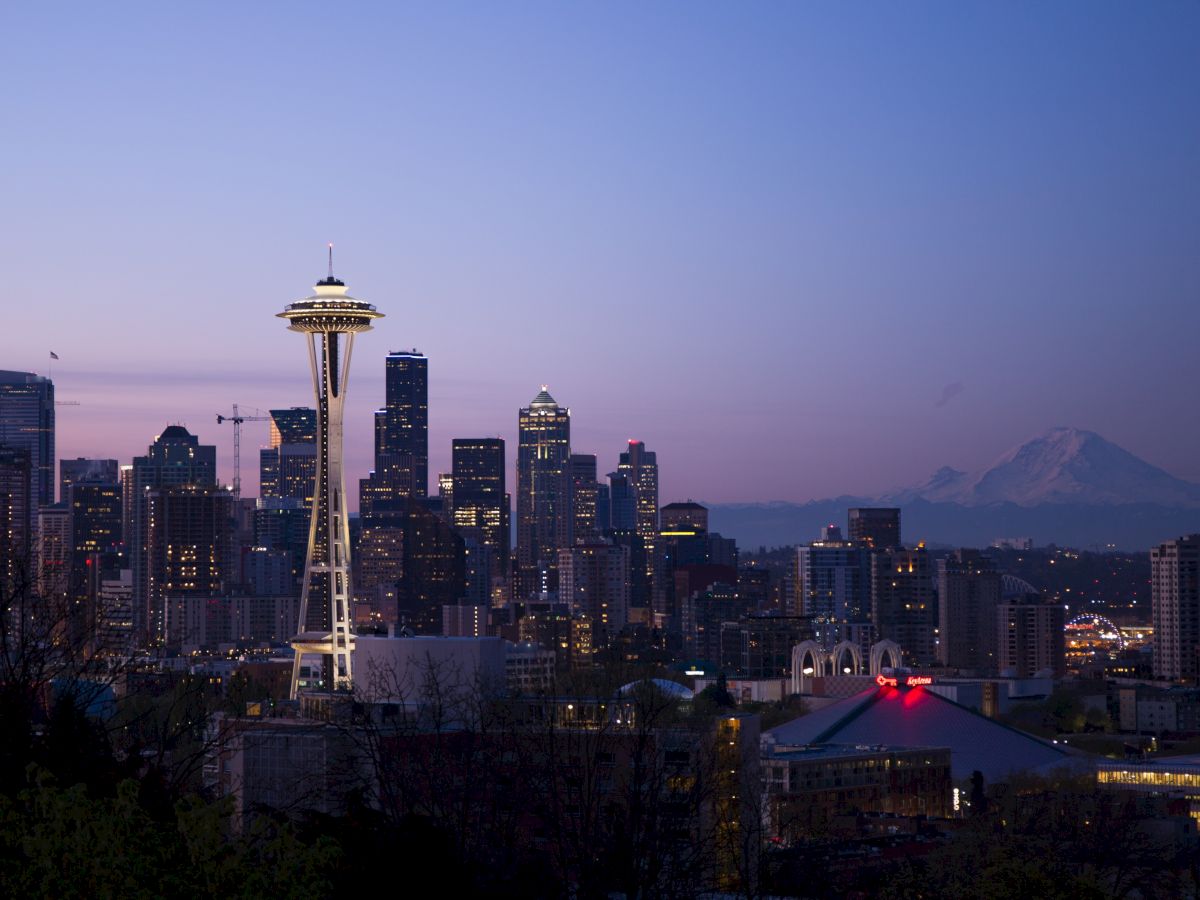 A cityscape featuring the Space Needle, tall buildings, and Mount Rainier in the background during twilight with clear skies ending the sentence.