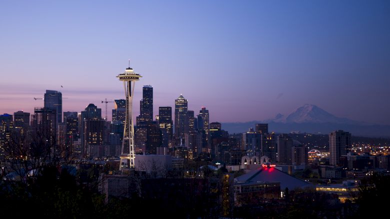 A cityscape featuring the Space Needle, tall buildings, and Mount Rainier in the background during twilight with clear skies ending the sentence.