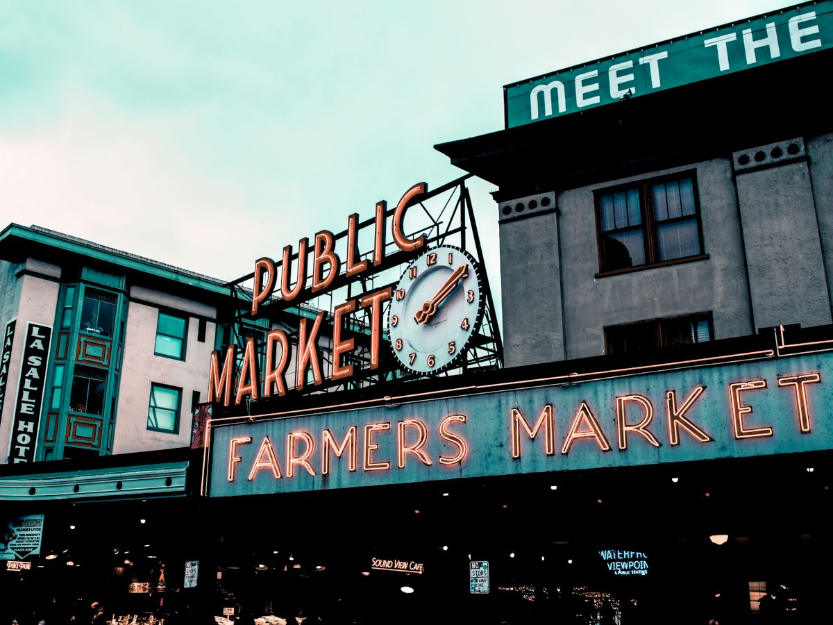 The image shows the entrance to a public farmers market, with neon signs and a clock. Buildings are visible in the background, including a hotel.