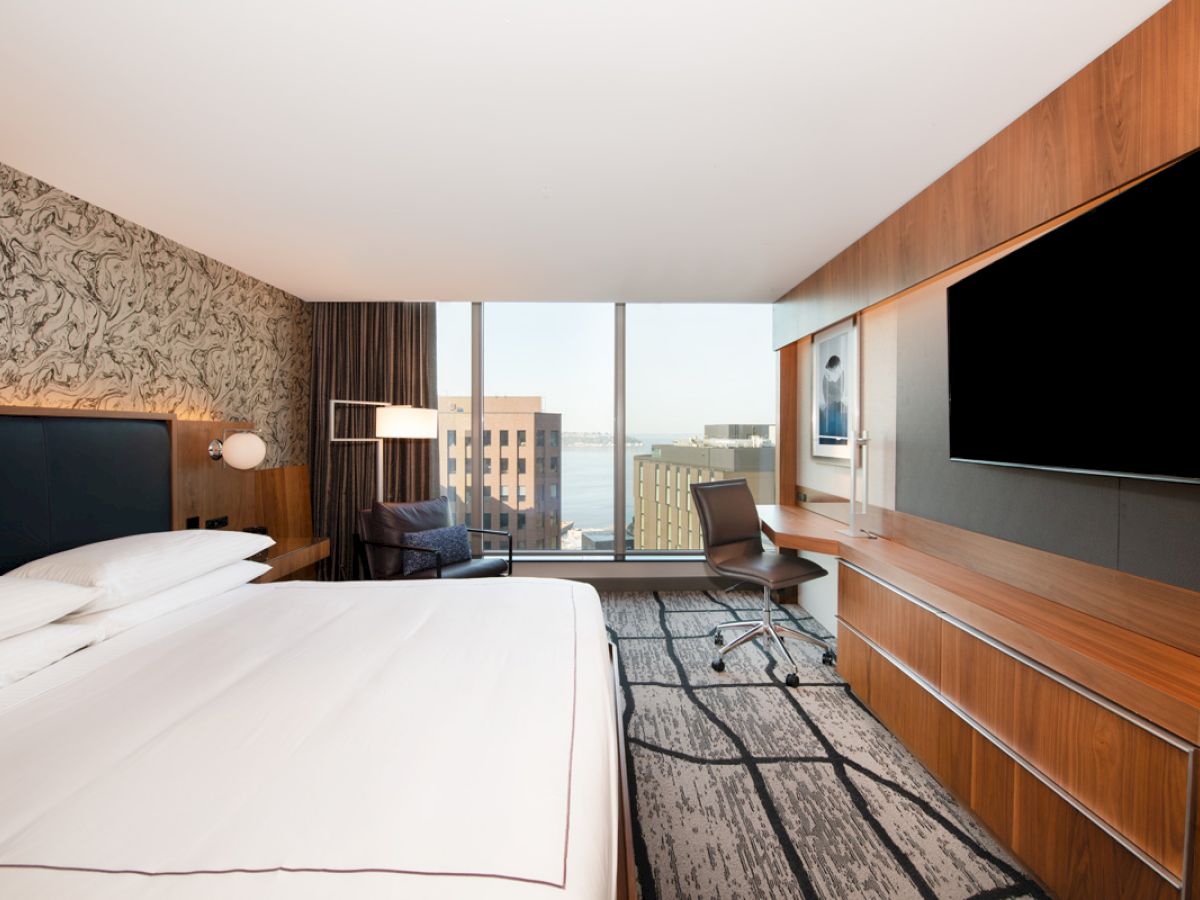 A modern hotel room featuring a large bed, a TV, a desk, and a chair near floor-to-ceiling windows with a city view.