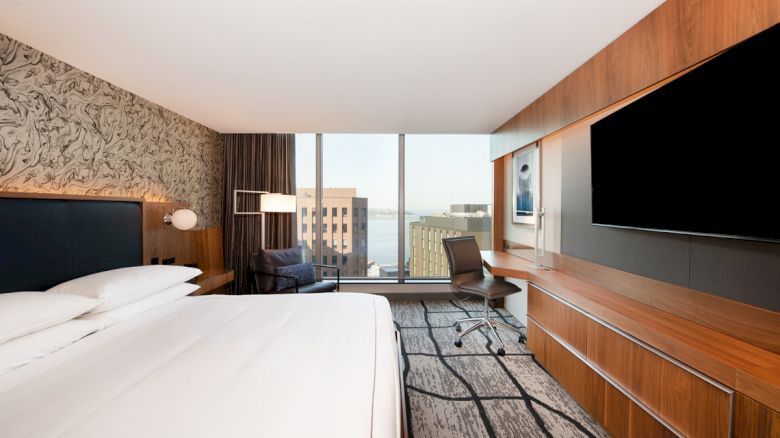 A modern hotel room featuring a large bed, a TV, a desk, and a chair near floor-to-ceiling windows with a city view.