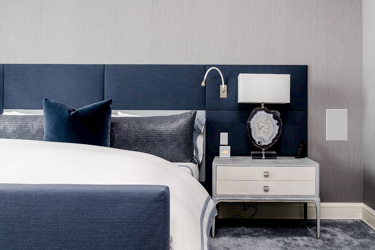A modern bedroom features a blue headboard, white bed linens, a nightstand with a lamp, a decoration, and a wall-mounted reading light.