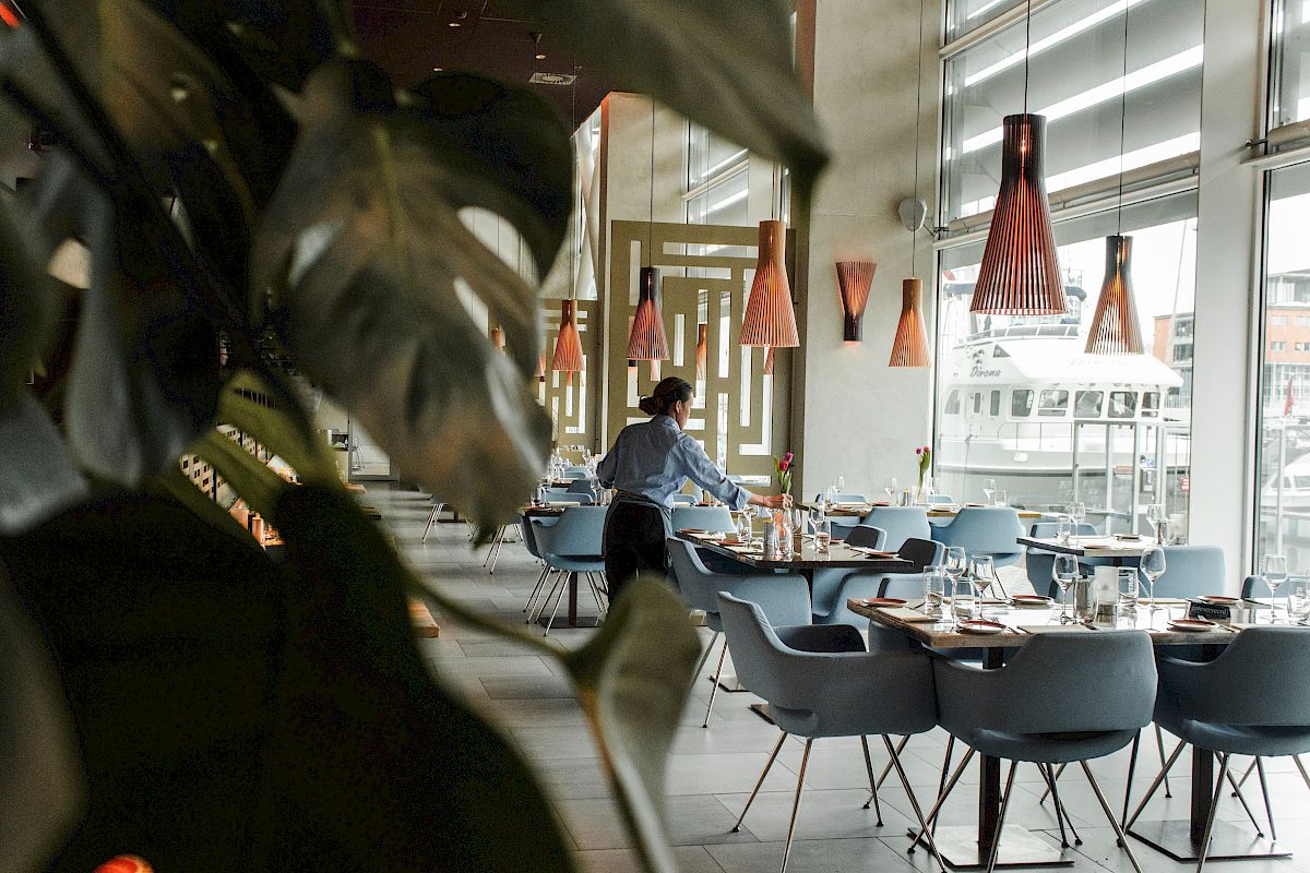 A person is arranging tables in a modern restaurant with stylish lighting, large windows, and plants, giving the space a cozy and elegant ambiance.