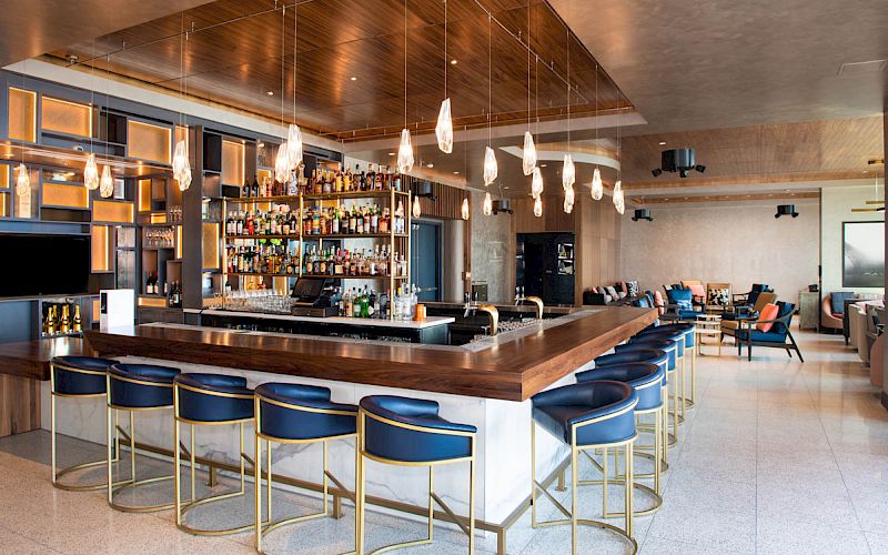 The image shows a modern bar with a wooden top, blue bar stools, bottle shelves, pendant lighting, and a seating area in the background.