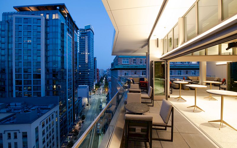 This image shows a modern balcony with outdoor seating, overlooking a cityscape with tall buildings during twilight hours.