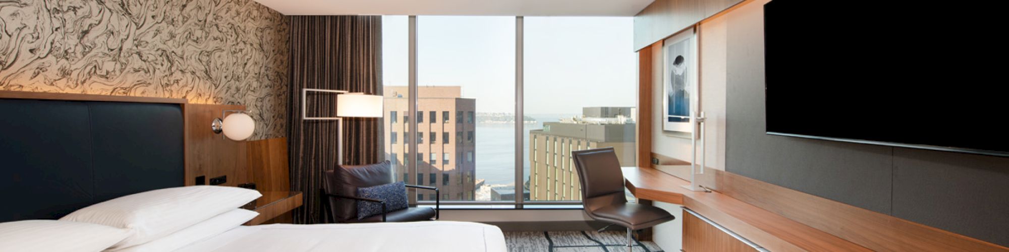 A modern hotel room features a large bed, wall-mounted TV, desk, chair, and window with a cityscape view, decorated in neutral tones.